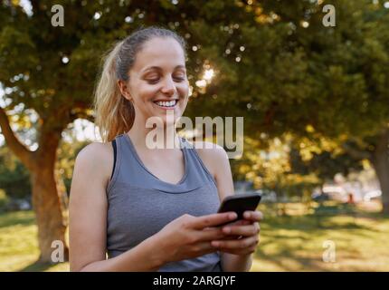 Portrait einer lächelnden, schönen blonden jungen Frau, die auf dem Smartphone im Park eine Nachricht getextet hat Stockfoto