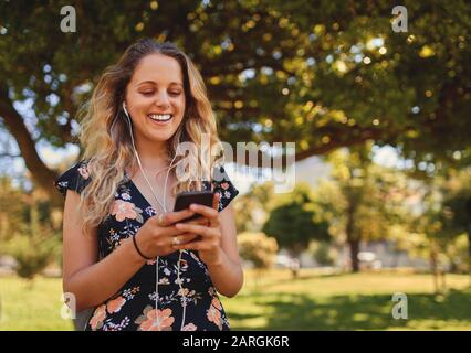 Porträt einer lächelnden, blonden jungen Frau mit Ohrhörern in den Ohren SMS auf ihrem Smartphone im Park an einem sonnigen Tag Stockfoto