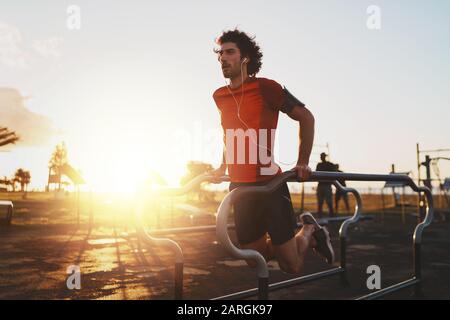 Sportlicher junger Athlet, der Musik auf Ohrhörern hört, die auf parallelen Bars trainieren, die Push-Ups im Park machen - junger Mann, der im Freien Dips macht Stockfoto