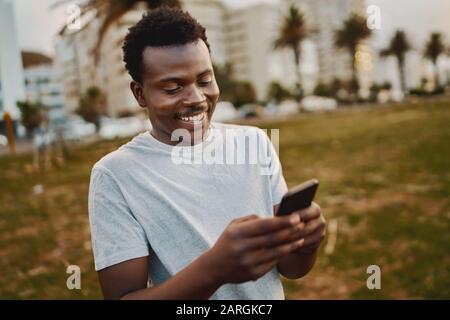 Porträt eines jungen männlichen Athleten, der lächelnd im Park steht, während er Nachrichten auf dem Handy textet
