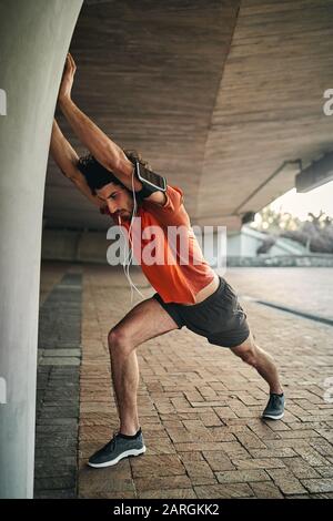 Gutaussehender junger Mann in Sportswear mit Ohrhörer und Armband, der sich beim morgendlichen Lauf aufwärmte, indem er Arm und Bein streckte Stockfoto