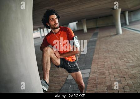 Porträt eines ernsthaften jungen Mannes, der sich das Bein unter der Brücke streckt, bevor er morgens joggt Stockfoto