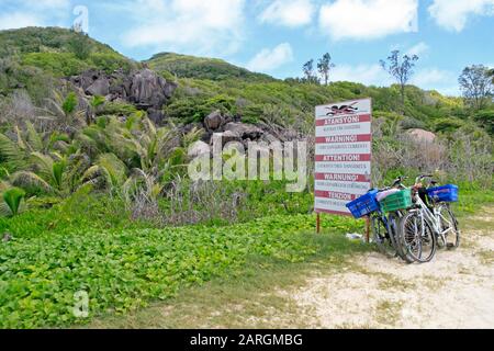 Fahrräder, die vor warnungsgefährlichen Strömungen geparkt wurden, signieren mit Wasserburschen und Palmen auf der Insel La Digue auf den Seychellen. Stockfoto