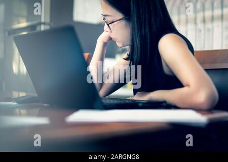 Geschäftsfrau sitzt am Tisch und liest Dokumente mit offenem Laptop. Stockfoto