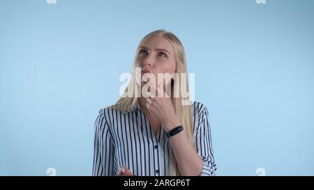 Blonde junge Frau hält ihr Kinn mit der Hand, während sie über etwas auf blauem Hintergrund nachdenkt. Sie sieht sich Smartphone an und macht Pläne. Stockfoto