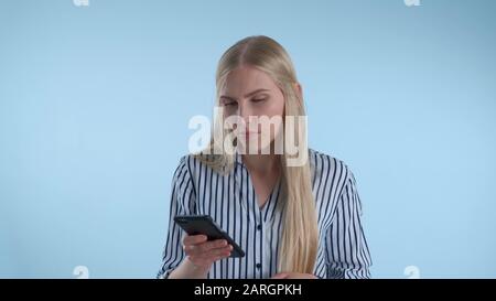 Blonde junge Frau hält ihr Kinn mit der Hand, während sie über etwas auf blauem Hintergrund nachdenkt. Sie sieht sich Smartphone an und macht Pläne. Stockfoto