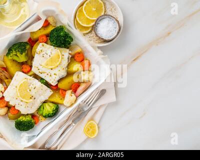 Fisch-Kabeljau im Ofen mit Gemüse gebacken - gesunde Ernährung gesundes Essen. Hellweißer Marmortisch, Kopierraum, Draufsicht Stockfoto
