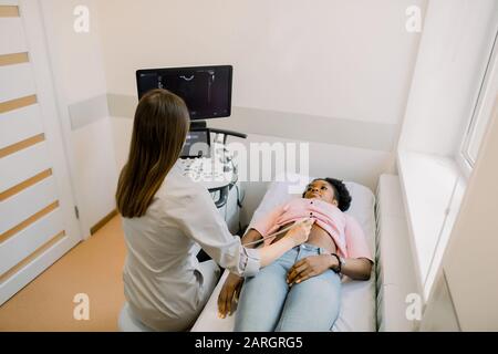 Ein junger Arzt betrachtet den Ultraschallbildschirm in einer modernen Klinik und führt gleichzeitig Ultraschall für junge Afrikanerin durch Stockfoto