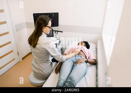 Glückliche junge afrikanische Patientin, die in der Klinik durch Abdomen-Ultraschall geht. Professioneller kaukasischer Frauenarzt, der Magenultraschall macht Stockfoto