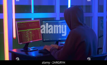 Mittelgroßer Hackerschuss, der in einer dunklen Nahaufnahme am Computer gekleidet ist. Er durchdringt das Netzwerksystem. Rückansicht Stockfoto