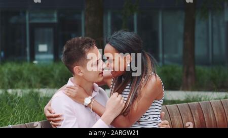 Zoom-Schuss des jungen Mixed-Race-Paares, das sich im Freien auf der Bank streichelt. Sie haben Datum in einem schönen Stadtpark. Stockfoto