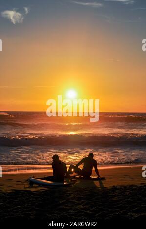 Silhouetten von zwei Surfern, die bei Sonnenuntergang am Strand sitzen Stockfoto