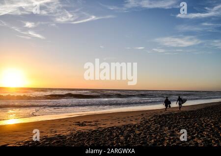 Silhouetten von zwei Surfern, die bei Sonnenuntergang am Strand spazieren Stockfoto