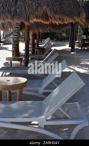 Weiße Lounge-Stühle in einer Linie unter Reetdachschirmen im Sand. Stockfoto