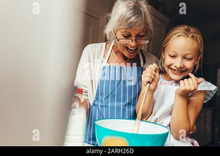 Glückliche Großmutter und Kind, die einen Kuchenteig zu Hause mischten. Fröhliches kleines Mädchen, das den Teig in einer Schüssel rührte, wobei ihre Granny an ihrer Seite stand. Stockfoto