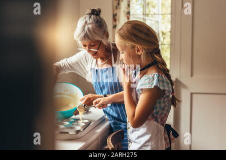 Großmutter unterrichte dem Kind, Tassenkuchen zu machen. Fröhliche Großmutter und Kind gießen Kuchenbatter in Tassenkuchformen. Stockfoto