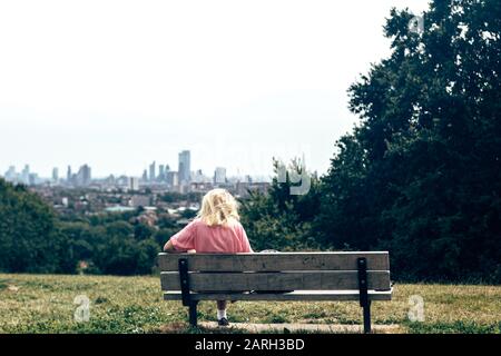 London/UK- 17/07/2019: Blonde Frau allein auf einer Bank auf dem Parliament Hill sitzend; Rückansicht. Parliament Hill ist eine Fläche offener Parklandschaft im Süden des Landes Stockfoto