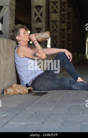 Eine Punkfrau unter einer Brücke trinkt Alkohol aus einer Flasche in einer braunen Papiertüte Stockfoto