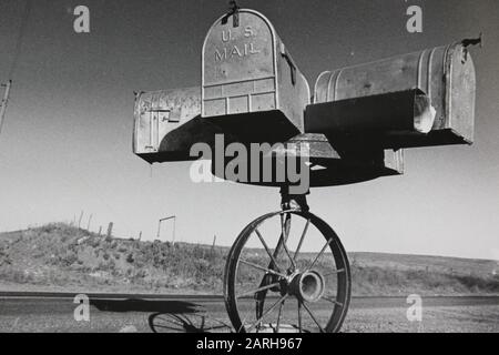 Feine Schwarzweißfotografie der 1970er Jahre einer US Mail Postbox auf einem Wagenrad auf den Ebenen Stockfoto