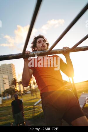 Jungen Mann mit Ohrhörern in die Ohren bringen und im Sommer seine Muskeln an Bars im Freien trainieren - Mann, der Pullups im Freien macht Stockfoto