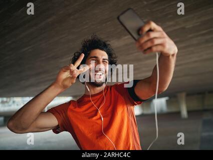 Portrait eines lächelnden jungen und gutaussehenden Mannes mit mobilem Smartphone, der ein Friedenszeichen zieht und ein selfie nimmt Stockfoto