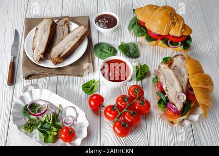 Nahaufnahme von Croissant Sandwiches mit zerschnittenen Schweinebauch, Tomaten, frischem Spinat, Zwiebel auf einem weißen Holztisch mit Zutaten auf einem Tisch, horizontal Stockfoto