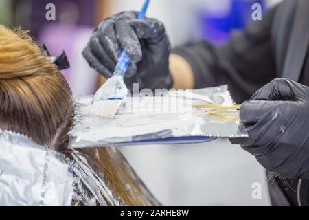 Hände eines Para-Friseursalons in schwarzen Handschuhen tragen eine Substanz zum Färben von Haaren auf einer Folie mit blauer Bürste auf Stockfoto