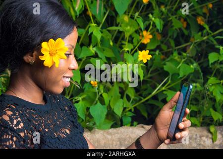 Schöne junge afrikanerin, die nachts ihr Handy im Freien benutzt, mit einer Blume im Haar Stockfoto