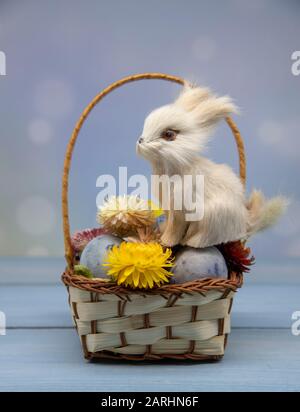 Osterhase sitzt in einem Korb mit bemalten Eiern und getrockneten Blumen auf einem Holztisch Stockfoto