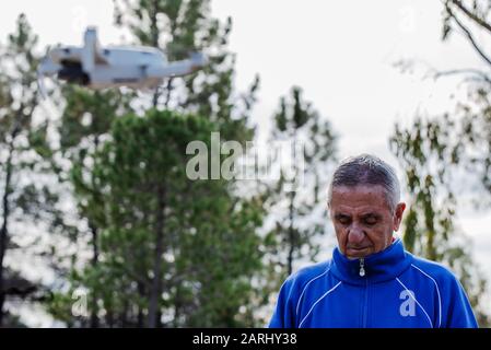 Die Seitenansicht des alten Mannes, der die Drohne mit Blick auf die Fernbedienung fliegt, während er im Park gegen die Bäume steht, konzentriert sich auf den alten Mann Stockfoto