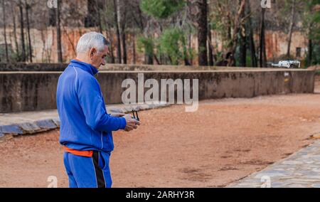 Seitenansicht des alten Mannes, der die Drohne in Sportswear fliegt, während er gegen die Bäume im Park steht Stockfoto