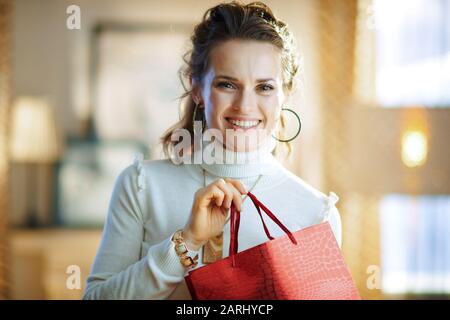 Porträt der fröhlichen Hausfrau im mittleren Alter in weißem Pullover und Rock mit roter Einkaufstasche im modernen Zuhause am sonnigen Wintertag. Stockfoto