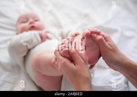 Baby feets in den Händen der Mutter. Die junge kaukasische Frau massiert zu Hause für glückliches Säuglingsbaby auf weißem Bett. Babypflege, Sport und glückliche Mutterschaft. Stockfoto
