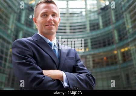 Lächelnder Geschäftsmann, der im Freien steht und im Innenhof eines modernen, geschwungenen Büroturms mit umgeklappten Armen umklappt ist Stockfoto