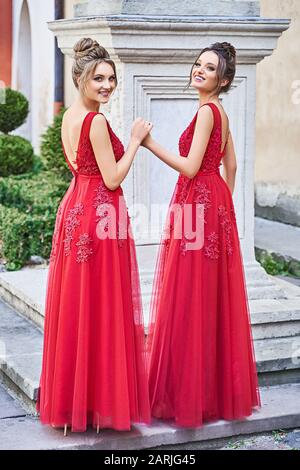 Zwei schöne Brautjungfer blonde und brünette Damen tragen ein elegantes rotes Chiffon Brautmädchenkleid mit Spitze und Holblume Stockfoto