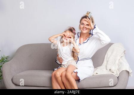 Fröhliche glückliche Mutter und Tochter mit Lockenwicklern im Haar, die in Ferngläsern mit den Fingern auf die Kamera schauen und zusammen auf dem Sofa lächeln, gr Stockfoto