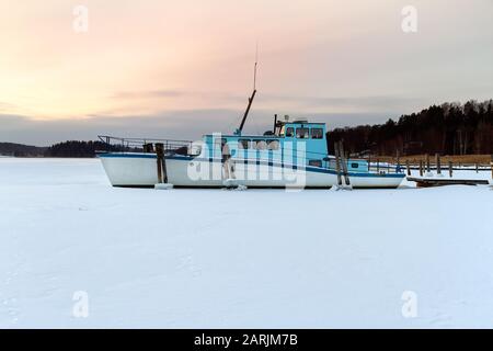 Das Boot ist im Winter im Eis stecken geblieben, Kaarina, Finnland. Schöner Sonnenuntergang im Hintergrund. Stockfoto