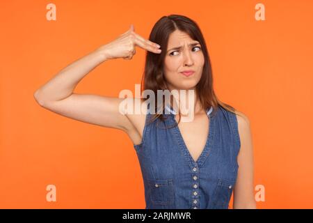 Verzweifelte brünette Frau im Denimkleid, die verängstigt auf Fingerpistole zeigte, die auf den Kopf zeigte und sich mit Selbstmordgeste tötete, erschrocken, lebensmüde. Stockfoto