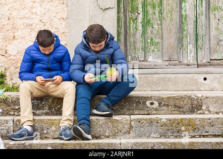 Italien, Sizilien, Provinz Messina, Montalbano Elicona. April 2019. Zwei Jungs spielen auf ihrem Handy, während sie auf palmsonntagsgottesdienste in Montal warten Stockfoto