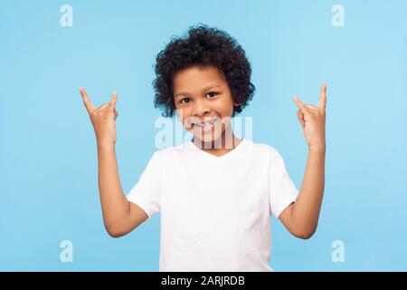 Lass uns rocken! Porträt eines fröhlichen funky kleinen Jungen mit lockigem Haar in weißem T-Shirt mit Rock- und Roll-Geste und lächelnd, das verrückte coole Symbol w Stockfoto
