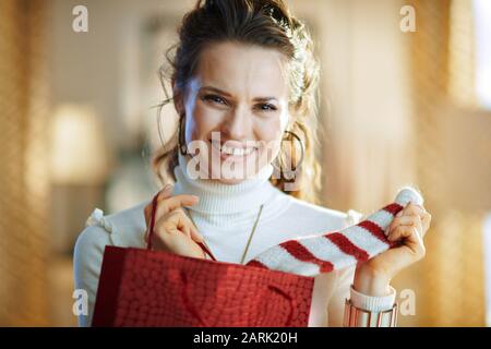 Porträt einer lächelnden eleganten Frau in weißem Pullover und Rock mit roter Einkaufstasche, die im modernen Wohnzimmer im sonnigen Wine gekauften Pullover auszieht Stockfoto