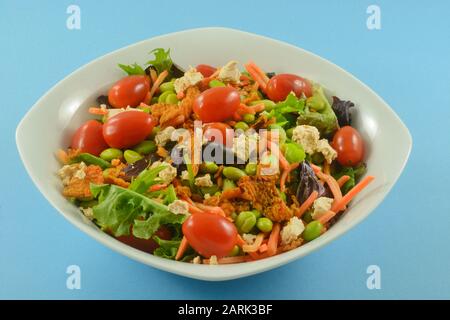 Gesunder Salat mit frischen gemischten Grüns, Traubentomaten, Streichholzkartoffeln, Edamame, Käse und knusprigen roten Paprika in weißer Schüssel auf blauem Grund Stockfoto