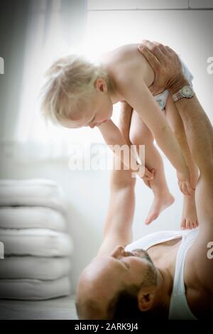 Kleines Kind in Windeln, das von seinem Vater, der auf dem Boden in einem Raum in ihrem Haus liegt, in der Höhe gehalten wird. Stockfoto