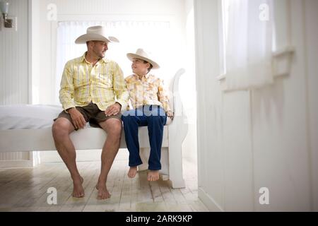 Reifer Mann sitzt neben seinem Sohn auf einem Bett in einem Haus, während beide Cowboy-Hüte tragen. Stockfoto