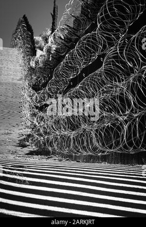 Blick auf den internationalen Grenzzaun USA/Mexiko mit Rasierdraht nach bergauf mit Mexiko auf der anderen Seite des Zauns von Nogales, AZ, USA, in B&W Stockfoto