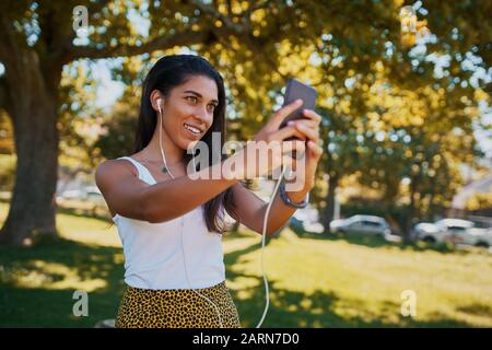 Lächelnde, trendige junge Frau mit Ohrhörern im Ohr, die selfie lächelnd und glücklich auf dem Smartphone im Park nimmt Stockfoto