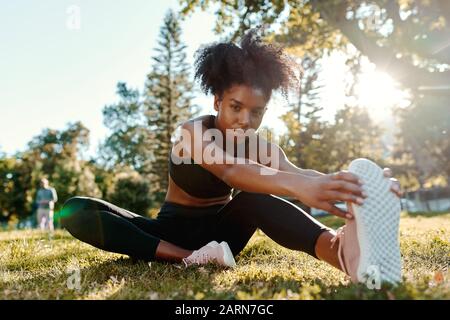 Portrait einer entschlossenen afroamerikanischen jungen Frau, die morgens auf grünem Gras sitzt, Sonnenlicht, die ihre Beine mit Blick auf die Kamera streckt