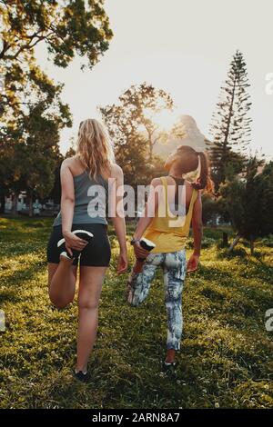 Rückansicht verschiedener multirassischer weiblicher Freunde, die sich im Sonnenlicht im Park die Beine Strecken Stockfoto