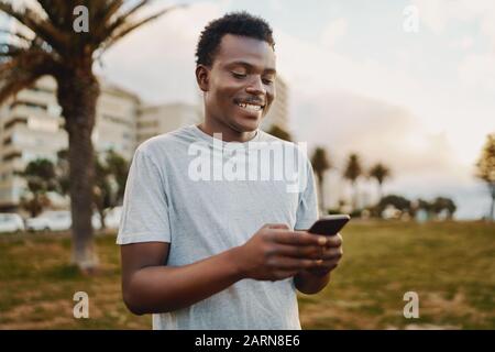 Portrait eines sportlichen jungen afroamerikanischen Mannes, der lächelt, während er Nachrichten auf dem Handy im Park textet Stockfoto