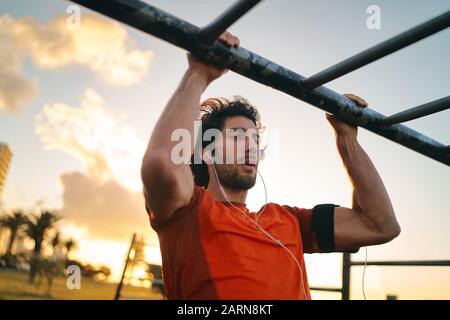 Gesunde Fitness junger Mann, der Musik auf Ohrhörern hört, die Pull-Ups im Sportpark im Freien machen - Mann, der Kin-Ups macht Stockfoto